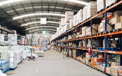 Warehouse storage facility interior. Large distribution center with shelves full of cardboard boxes, pallet jack machine, and package drinking water bottle stacks.