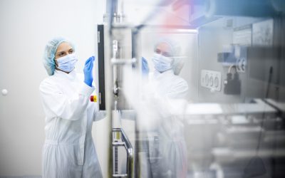 A woman wearing proper safety clothes seen taping on the screen of a big complex machine in pharmaceutical industry while her reflection is visible on the machine.