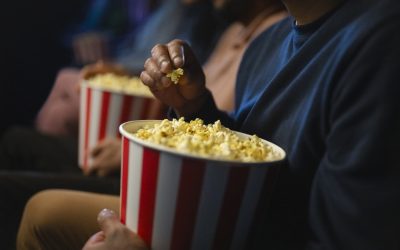 A close-up shot of a hand grabbing popcorn from a red and white striped container, symbolizing the enjoyment of a movie theater experience.