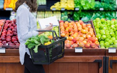 Young woman carries a shopping basket filled with fresh produce. She is shopping for fresh fruit and vegetables in a grocery store.
